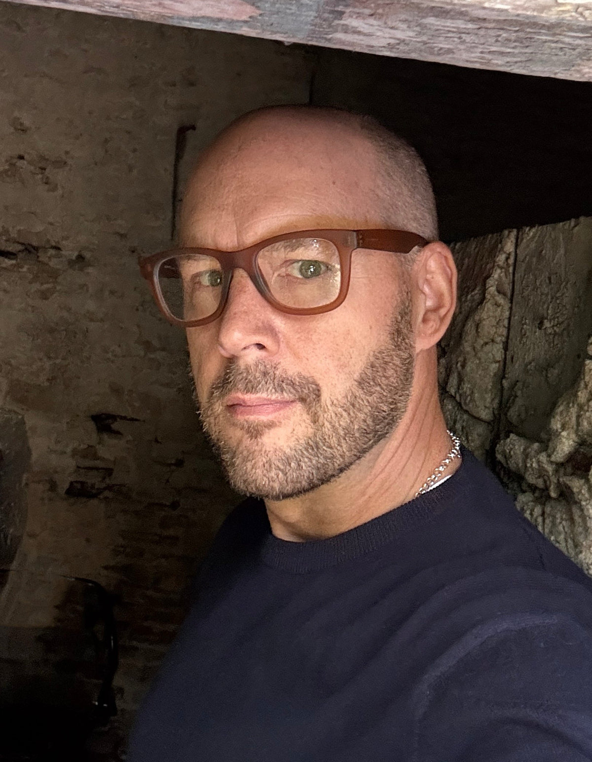 A close-up portrait of a man with a shaved head and beard wearing brown honey-rimmed Italia Independent eyeglasses and a dark navy crewneck sweater, photographed in a dim, rustic stone or wood interior.