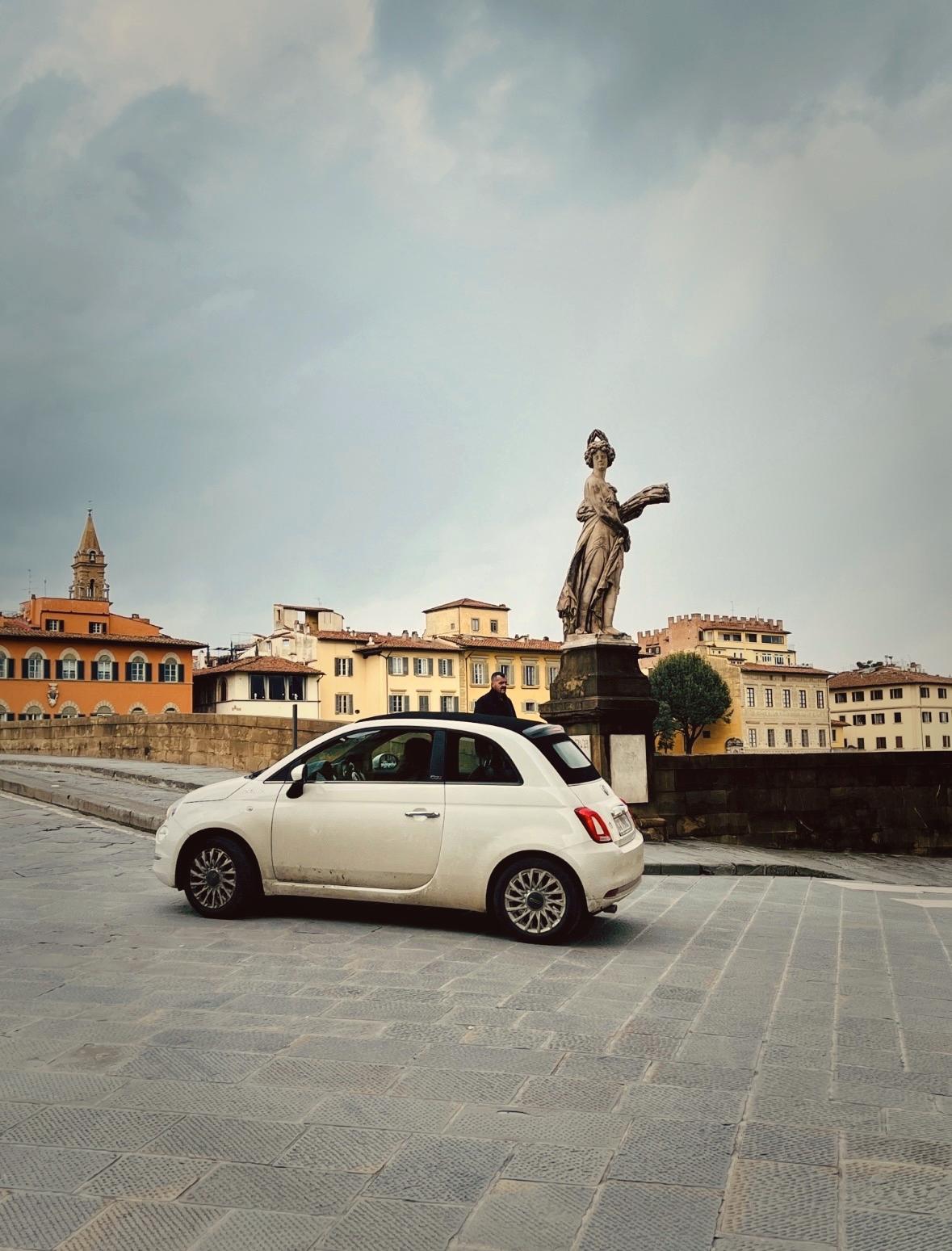 White FIAT 500 car parked on a stone pavement with a statue and buildings in the background in Florence Italy