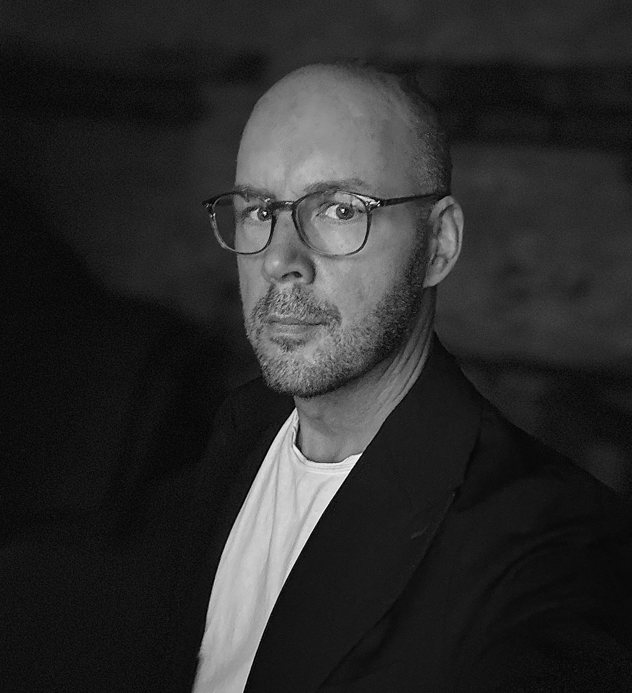 A black and white portrait of a man with a shaved head, wearing panto eyeglasses, a white t-shirt, and a dark  blue blazer.